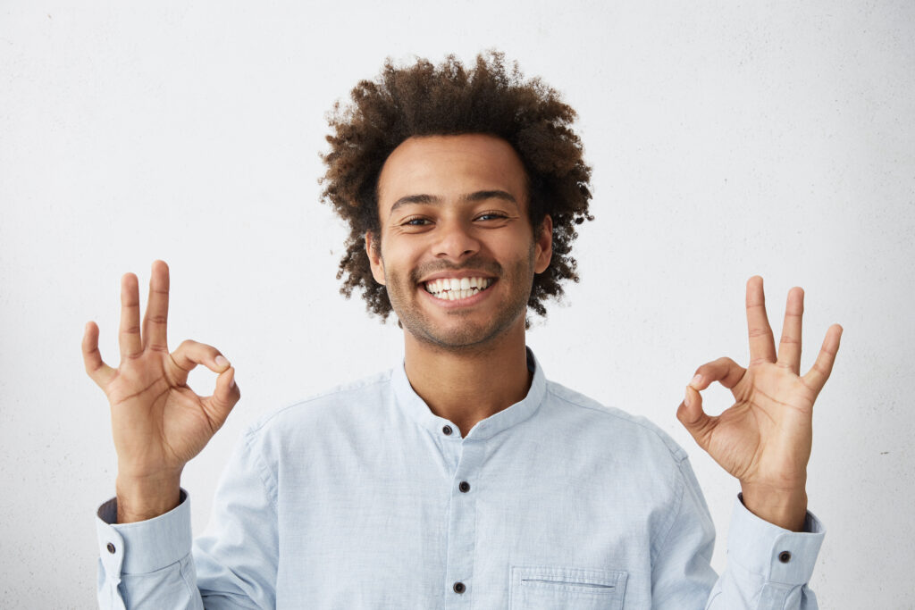 Positive human facial expressions, emotions and feelings. Carefree happy young African American man with funky hairstyle showing Ok sign with both hands and smiling broadly, enjoying good nice day